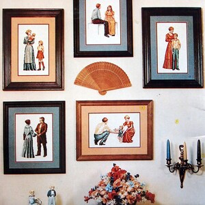 Puede incluir: Cinco dise&ntilde;os de bordado de punto de cruz enmarcados que representan familias en varias poses. Los marcos son de madera marr&oacute;n con un borde dorado. Un abanico de madera est&aacute; colgado en la pared sobre el marco central. Un arreglo floral est&aacute; sobre una mesa en primer plano.
