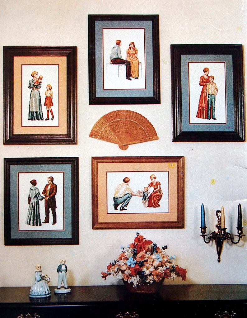 Puede incluir: Cinco dise&ntilde;os de bordado de punto de cruz enmarcados que representan familias en varias poses. Los marcos son de madera marr&oacute;n con un borde dorado. Un abanico de madera est&aacute; colgado en la pared sobre el marco central. Un arreglo floral est&aacute; sobre una mesa en primer plano.