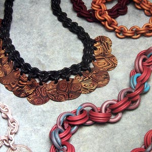 Puede incluir: Una colección de joyas que incluye un collar de cadena negra con colgantes circulares veteados en marrón y negro, una cadena de eslabones roja y azul, una cadena roja oscura, una cadena naranja y una cadena rosa pálido con un colgante.