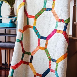 May include: A white quilt with a geometric pattern of colorful, connected hexagons. The quilt features a variety of fabrics in shades of red, orange, yellow, green, blue, and pink. The background is a quilted white fabric.