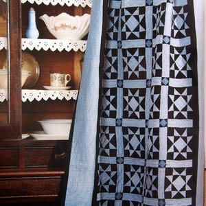 May include: A handmade quilt with a geometric Ohio Star pattern in black and light blue. The quilt is draped over a dark wooden cabinet with white shelves displaying various ceramic items. The quilt's border is light blue.