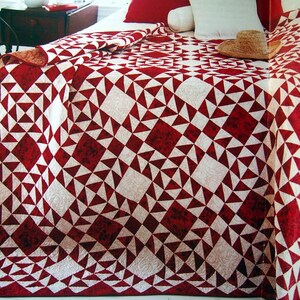 May include: A red and white quilt with a geometric pattern of triangles and squares. The quilt is spread out on a bed, with a straw hat resting on top. The quilt's design features a repeating pattern of red and white shapes.