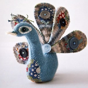 May include: Handmade blue fabric peacock with button and fabric feather details. The peacock has a light blue body, a white beak, and a crown of green thread. The tail feathers are a mix of patterned fabrics and buttons.