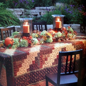 May include: An outdoor table setting with a quilted tablecloth in brown, red, and beige. Two glass candle holders with lit orange candles are the centerpiece, surrounded by pumpkins and autumn leaves. Black chairs are arranged around the table, creating a warm atmosphere.