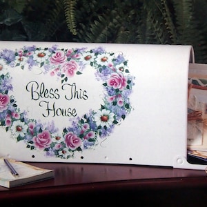 Puede incluir: Buzón de metal blanco con un diseño de corona floral pintada en tonos de rosa, morado y blanco. La corona rodea las palabras "Bless This House".