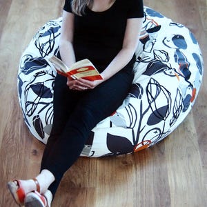 May include: A large, round beanbag chair with a white background and a black, gray, and orange floral pattern. The chair is on a wooden floor. The person is wearing black clothing and orange and brown sandals.