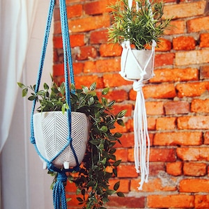May include: Two hanging planters with greenery against a brick wall. One planter is white with a black chevron pattern and blue rope. The other is a white planter with white rope. Both planters hold green plants.