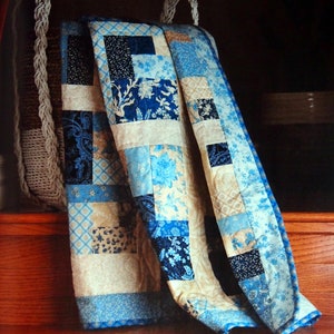 May include: A blue and cream patchwork quilt with a floral pattern is draped over a woven basket. The quilt is folded and has a blue border.