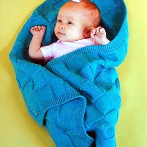 Könnte beinhalten: Ein Baby, das in eine türkisfarbene gestrickte Decke mit Rautenmuster gewickelt ist. Das Baby trägt ein rosa Hemd und hat eine weiße Schleife im Haar.
