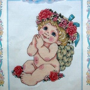 Puede incluir: Punto de cruz de una figura querubínica con rizos rubios, ojos azules y una corona floral. La figura está sentada, con las manos juntas en oración, rodeada de rosas rojas y pequeñas flores. El fondo es azul claro.