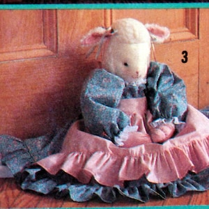 May include: A white lamb doll wearing a pink and blue dress with a ruffled skirt. The doll is sitting by a wooden door with the number "3" on it.