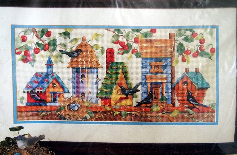 May include: Cross-stitch pattern featuring a row of colorful birdhouses with a cherry tree and birds. The birdhouses are decorated with different patterns and colors, including blue, green, red, and yellow. The cherry tree has red cherries and green leaves. The birds are black and have yellow beaks.