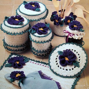 May include: A collection of handmade crochet kitchen accessories, including canisters, a utensil holder, a jar cover, a placemat, and a napkin ring. Each item is white with teal trim and a purple flower with a yellow center.
