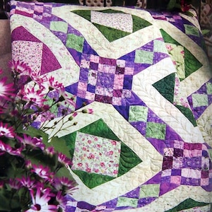 May include: A quilt with a diamond pattern in shades of purple, green, and white. The quilt is made with floral fabric and has a quilted design.