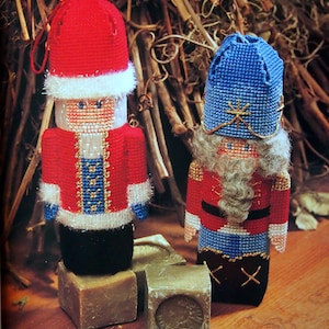 May include: Two handmade Christmas ornaments, one red and white and one blue and white, depicting a Santa Claus and a Nutcracker. Both ornaments are made with cross-stitch and have a 3D effect.