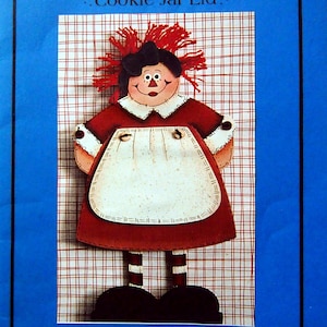 May include: A red and white Raggedy Annie cookie jar lid craft pattern. The pattern features a smiling Raggedy Annie doll with red hair, a white apron, and black shoes. The pattern is designed by Trisha Biesinger and is exclusively for Macs Hobby & Craft Superstores.