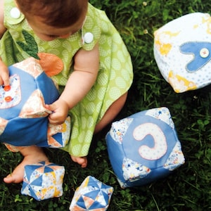 May include: A baby plays with colorful fabric blocks on a green grassy surface. The blocks are decorated with letters and patterns.