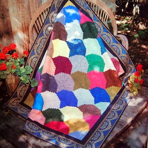 Op de afbeelding: Een kleurrijk quilt met een blauwe, gouden en zwarte rand. De quilt is gemaakt van verschillende stoffen in tinten rood, groen, blauw, geel en bruin. De quilt is gevouwen en over een houten stoel gelegd.