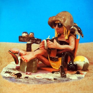 May include: A whimsical figurine depicting a person relaxing on a beach chair, wearing sunglasses and a hat. The figurine includes a beach ball, camera, and a sign that reads "DO NOT DISTURB". The background is blue, with sand and a small bird.