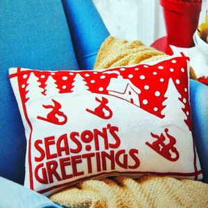 May include: A white pillow with red polka dots and a red embroidered design of a winter scene with the text "Season's Greetings". The pillow is on a blue armchair with a tan blanket.