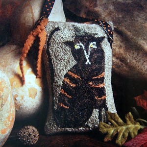 May include: A black cat with orange stripes is depicted on a gray and white fabric wall hanging. The cat has yellow eyes and a white nose. The wall hanging has a brown fringe and a black and white polka dot strap.