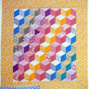May include: A quilt with a geometric pattern of colorful cubes. The cubes are made of different fabrics with floral prints. The quilt is surrounded by a yellow border with a floral pattern.
