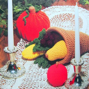 Op de afbeelding: Gehaakte herfstdecoraties, waaronder een pompoen, een hoorn des overvloeds en een appel. De decoraties zijn op een wit gehaakt tafellaken met twee zilveren kandelaars en witte kaarsen.