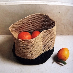 May include: A woven basket with a black base and tan upper section, holding three oranges. One orange rests outside the basket, next to a green leaf. The basket has a wavy design.