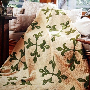 May include: A beige quilt with a repeating green leaf pattern. The quilt is draped over a window seat with pillows. The leaf design is a stylized floral pattern. The quilt appears to be handmade and has a quilted texture.