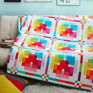 Op de afbeelding: Een kleurrijk patchwork-deken met een geometrisch ontwerp in tinten rood, oranje, geel, groen, blauw en paars. Het deken is gevouwen en tentoongesteld op een witte bank.