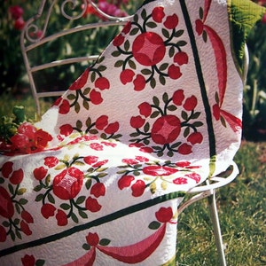 May include: A white quilt with a red and green floral pattern. The quilt is folded and draped over a white metal chair. The chair is sitting on a green grassy lawn.