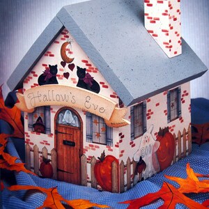 Puede incluir: Una casa de madera decorativa de Halloween con techo gris y chimenea. La casa presenta detalles pintados que incluyen gatos negros, calabazas, un fantasma y el texto "Hallow's Eve" en una pancarta. La casa está rodeada por una valla blanca.
