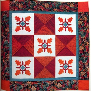 May include: A quilt with a white background and a pattern of red, orange, and teal squares. The squares are arranged in a grid pattern and each square has a different design. The quilt is surrounded by a border of dark green and orange leaves.
