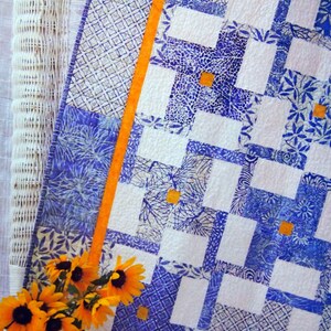 May include: A blue and white quilt with a geometric pattern and orange trim. The quilt is made of fabric squares with different designs. The quilt is on a white wicker chair with a bouquet of yellow flowers in front of it.