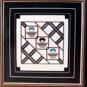 May include: Framed cross-stitch artwork with three square floral designs in teal and brown. The squares are set against a white background with a black and white crisscross pattern. The frame is gold and black.