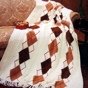 May include: A white crocheted blanket with brown and orange diamond patterns. The blanket is draped over a brown couch and has fringe on the bottom edge. The text "Bernat" is visible in the upper right corner of the image. The text "Book No. 511 Featuring Berella 4* Yarn Price $2.00 In Canada: $2.40" is visible in the upper left corner of the image.