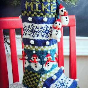 May include: A handmade blue and white Christmas stocking with the name "MIKE" knitted in green. The stocking features a snowman design and white pom-poms. A red chair and gifts are in the background.
