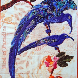 May include: A fabric art piece featuring two blue birds perched on branches, with a flower and the text "Bird of Paradise." The birds have detailed wing patterns in shades of blue, green, and purple. The background is a light, patterned fabric.