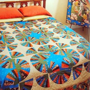 May include: A beige quilt with a fan-shaped pattern in shades of blue, green, orange, and brown. The quilt is on a bed with two pillows and a wooden headboard. A framed print with a Japanese scene is leaning against the wall.