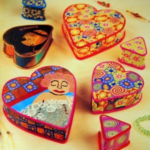 May include: A collection of colorful, heart-shaped and triangular decorative boxes. The boxes feature intricate patterns, including floral and geometric designs, and a painted image of a woman in a bath. The boxes are trimmed in red and pink.