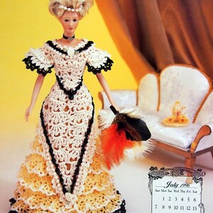 May include: A doll in a cream and yellow crocheted dress with black edging, holding a feather fan. The doll has blonde hair and a pearl headband. A calendar for July 1996 is visible in the background.