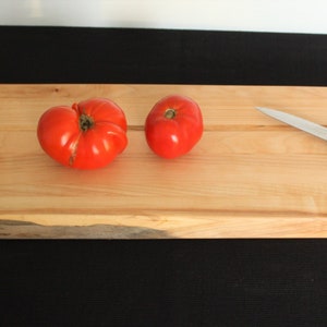 Peut inclure: Une planche à découper en bois rectangulaire et longue, avec une finition en bois naturel. La planche a un bord brut et deux rainures qui courent dans le sens de la longueur. Deux tomates rouges et un petit couteau argenté avec un manche noir sont sur la planche.