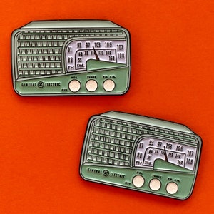 May include: Two green and white enamel pins shaped like vintage radios. The pins feature a dial with numbers and a knob. The text "GENERAL ELECTRIC" is printed on the bottom of each pin.