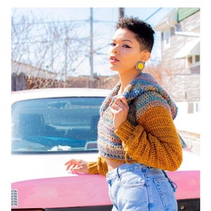 May include: A woman wearing a multi-colored crochet hoodie with a brown crocheted sleeve. The hoodie is cropped and has a hood. She is standing in front of a pink car. The text "HARMONY HOODIE" and "crochet pattern" are visible below the image. The text "serendipity-as-always.com" is visible below the text.