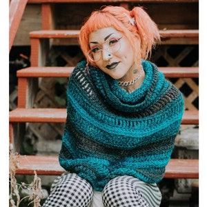 May include: A teal and gray crochet shawl with a cowl neck. The shawl is worn by a person with pink hair and glasses. The person is sitting on wooden steps.