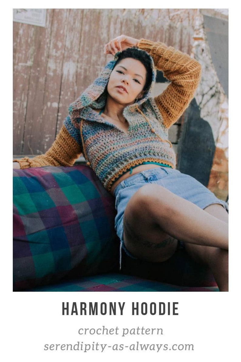 May include: A woman wearing a colorful crochet hooded top with brown sleeves and denim shorts. The top has a striped pattern and is cropped. The woman is sitting on a plaid cushion.