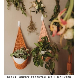 Puede incluir: Dos maceteros colgantes de crochet en naranja y rosa cuelgan de una pared con un gancho blanco. Los maceteros est&aacute;n llenos de plantas en maceta.