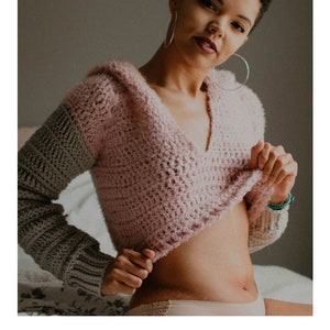 May include: A pink and gray crochet hoodie with a hood. The hoodie is cropped and has a V-neck. The text "HARMONY HOODIE" and "crochet pattern" are visible below the image. The text "serendipity-as-always.com" is visible below the text.