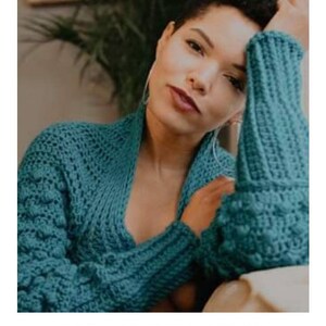 Op de afbeelding: Een tealblauwe gehaakte sjaal met een textuurpatroon. De sjaal wordt gedragen door een persoon met kort, krullend haar. De tekst "STELLAR MOON SHRUG" en "crochet pattern" is zichtbaar onder de afbeelding. Het websiteadres "serendipity-as-always.com" is ook zichtbaar.