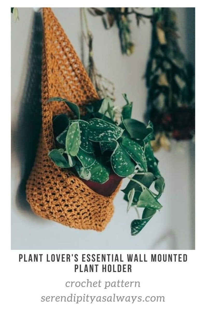 Puede incluir: Un macetero de crochet de color marr&oacute;n c&aacute;lido cuelga de una pared blanca. El macetero tiene forma de tri&aacute;ngulo y contiene una planta con hojas verdes. El texto "PLANT LOVER'S ESSENTIAL WALL MOUNTED PLANT HOLDER crochet pattern  serendipityasalways.com" est&aacute; debajo de la imagen.